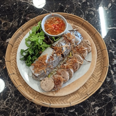 A woven bamboo tray contains a plate of Asian cuisine with fresh vegetables, steamed rolls topped with crispy fried onions, and sliced sausages arranged neatly. A small bowl of dipping sauce with red chilies sits next to the main dish.