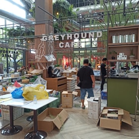 A cafe interior with a sign reading 'Greyhound Cafe'. There are people working, unpacking or arranging items. Several cardboard boxes, some open and filled with packing material or products, are scattered around. Tables hold bags and various objects, suggesting a busy or transitional state. The cafe is decorated with greenery and has a modern industrial design with exposed ducts and metal framework.