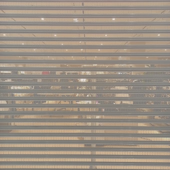 A closed metal security shutter with horizontal slats covers a storefront. Behind the shutter, interior lighting reveals an organized but slightly obscured view of a retail store with clothing racks and shelves visible.