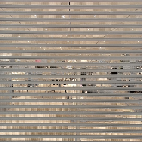 A closed metal security shutter with horizontal slats covers a storefront. Behind the shutter, interior lighting reveals an organized but slightly obscured view of a retail store with clothing racks and shelves visible.