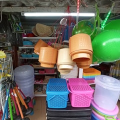 A diverse collection of household items and containers is displayed inside a shop. Hanging baskets in shades of beige and brown are suspended by red strings. Bright green inflatable balls are visible to the side. Stacked plastic baskets in blue, pink, and black, along with clear storage bins, are arranged below. Various other products, including hats and plastic tools, are neatly organized on shelves.
