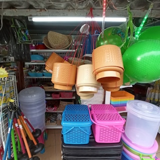 A diverse collection of household items and containers is displayed inside a shop. Hanging baskets in shades of beige and brown are suspended by red strings. Bright green inflatable balls are visible to the side. Stacked plastic baskets in blue, pink, and black, along with clear storage bins, are arranged below. Various other products, including hats and plastic tools, are neatly organized on shelves.