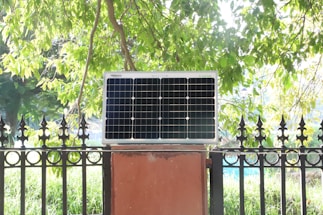 A solar panel is mounted on a red brick platform against a backdrop of lush green foliage. The sunlight filters through the tree leaves, casting a bright, natural light on the scene.