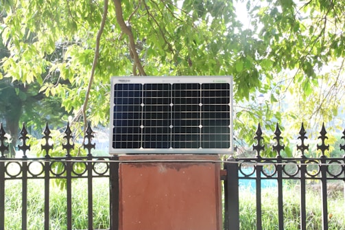 A solar panel is mounted on a red brick platform against a backdrop of lush green foliage. The sunlight filters through the tree leaves, casting a bright, natural light on the scene.