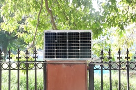 A solar panel is mounted on a red brick platform against a backdrop of lush green foliage. The sunlight filters through the tree leaves, casting a bright, natural light on the scene.