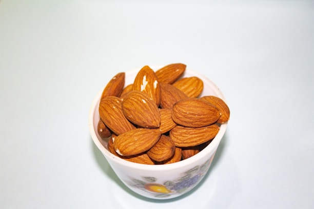 A white cup filled with whole almonds placed against a light blue background. The almonds are richly brown and have a smooth, natural texture.