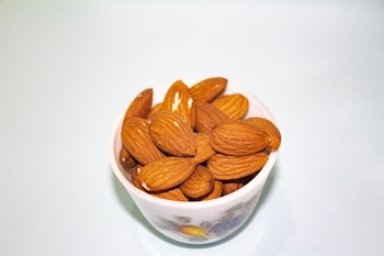 A white cup filled with whole almonds placed against a light blue background. The almonds are richly brown and have a smooth, natural texture.