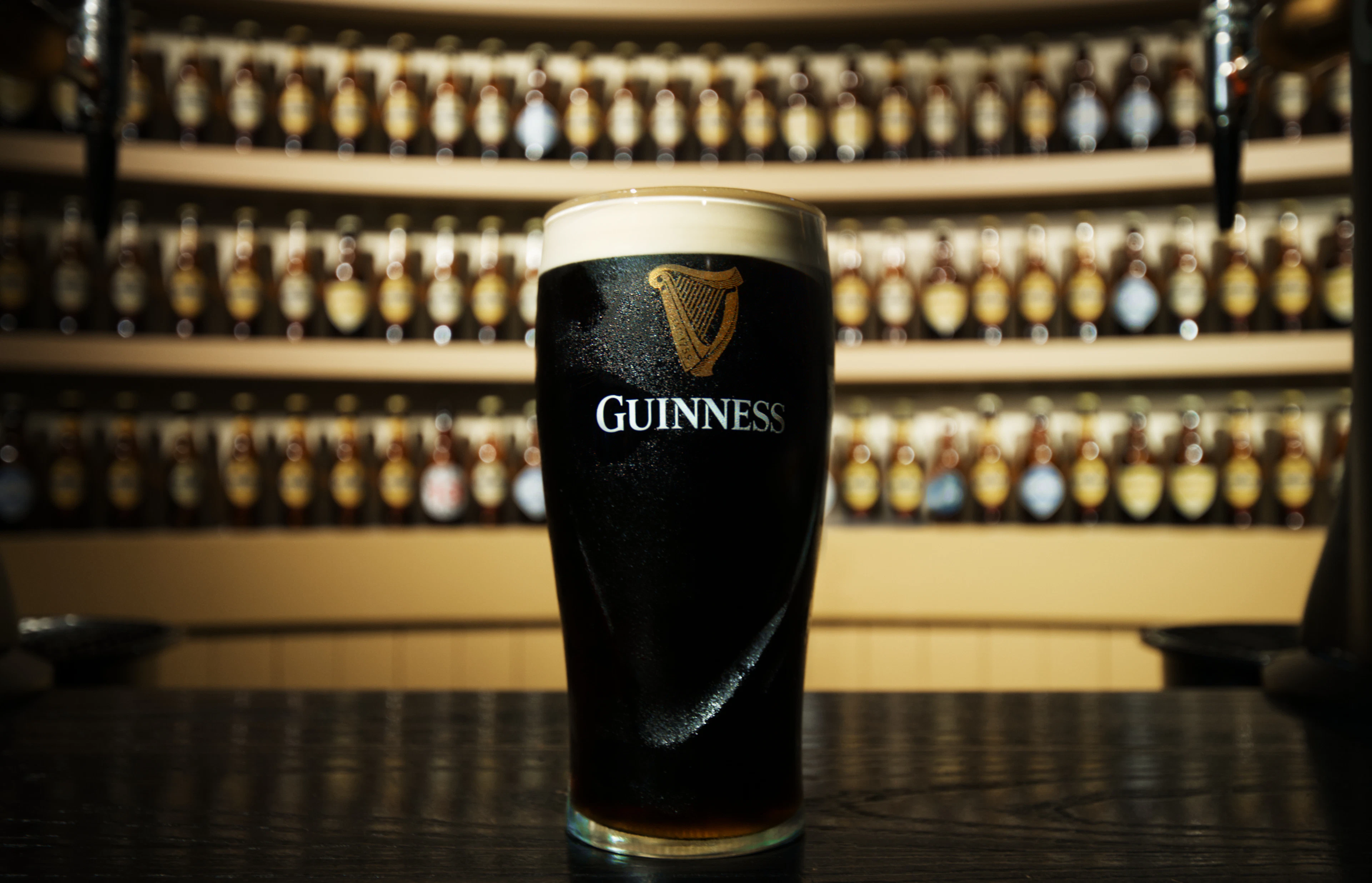A pint of Guinness stout with a thick creamy head standing on a bar counter in Dublin, Ireland, backlit rows of bottles behind it