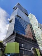 A modern skyscraper with reflective glass panels and angular architectural features dominates the scene. The words 'Global Trade Square' are displayed on a sign at the base of the building. A second, similarly styled building can be seen in the background. The clear blue sky enhances the urban setting.