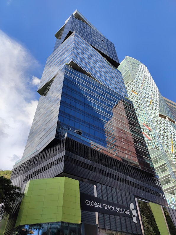 A modern skyscraper with reflective glass panels and angular architectural features dominates the scene. The words 'Global Trade Square' are displayed on a sign at the base of the building. A second, similarly styled building can be seen in the background. The clear blue sky enhances the urban setting.