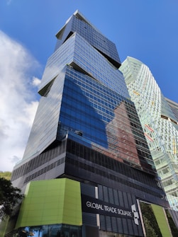 A modern skyscraper with reflective glass panels and angular architectural features dominates the scene. The words 'Global Trade Square' are displayed on a sign at the base of the building. A second, similarly styled building can be seen in the background. The clear blue sky enhances the urban setting.