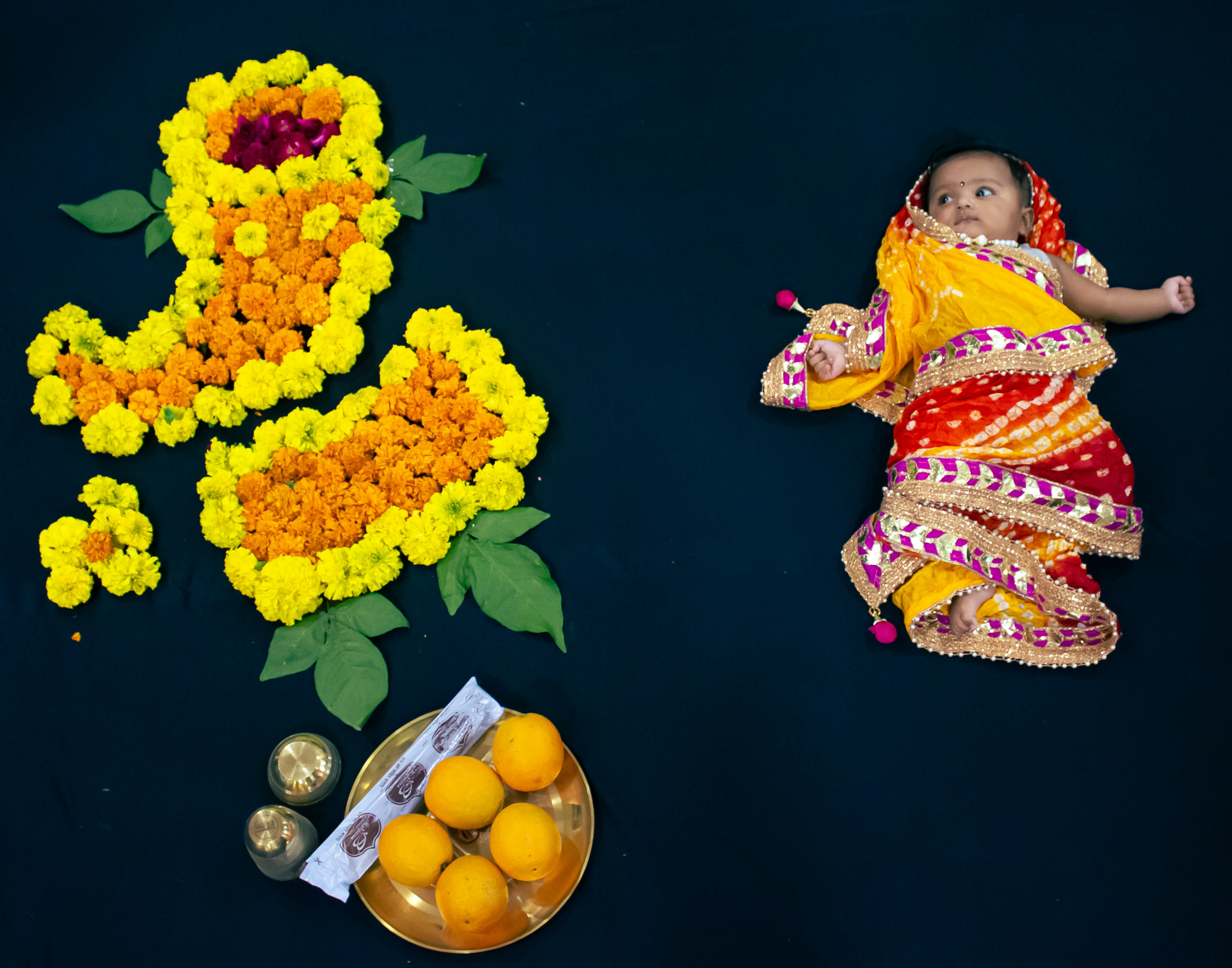 a baby dressed in a colorful outfit next to flowers and a plate of oranges