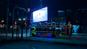 A nighttime urban street scene featuring a truck transporting a large number of motorbikes. The area is dimly lit with streetlights, and there's a bright billboard in the background displaying an advertisement. The atmosphere appears quiet and somewhat desolate.