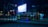 A nighttime urban street scene featuring a truck transporting a large number of motorbikes. The area is dimly lit with streetlights, and there's a bright billboard in the background displaying an advertisement. The atmosphere appears quiet and somewhat desolate.