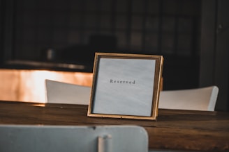 A wooden table with a framed sign displaying the word 'Reserved'. The sign is positioned in the center, and the background is softly blurred, suggesting a dimly lit indoor setting.