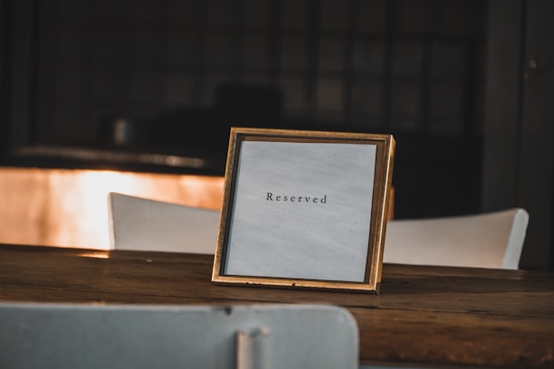 A wooden table with a framed sign displaying the word 'Reserved'. The sign is positioned in the center, and the background is softly blurred, suggesting a dimly lit indoor setting.