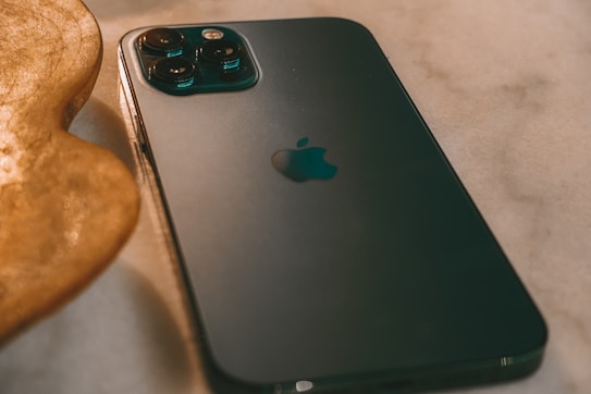 A smartphone with a sleek, dark design is placed on a smooth surface, with its camera lenses prominently visible. The iconic logo is centered on the back. The lighting creates a warm ambiance with shadows and reflections.