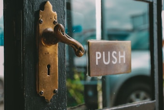 A vintage brass door handle and keyhole on a dark wooden door, alongside a rectangular metal push plate. A blurred reflection of a vehicle can be seen through a glass pane next to the door.