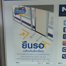 A safety poster featuring an illustration of a train platform with passengers inside a train. Arrows guide commuters to stand behind the yellow line. The poster includes both Thai and English text, emphasizing safety instructions for standing back until passengers have disembarked from the train.