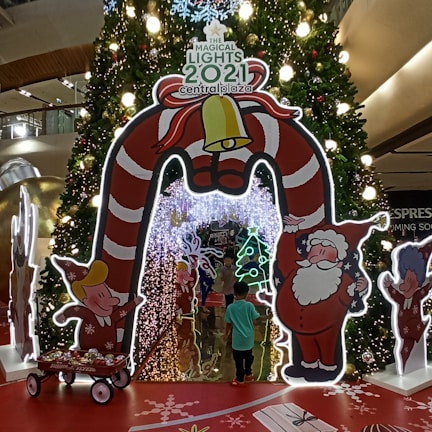 A festive holiday display featuring a large Christmas tree adorned with lights and ornaments. At the forefront is a decorative archway shaped like candy canes and a bell, with cartoon figures resembling elves and Santa Claus. A child is walking through the archway, surrounded by colorful decorations including a small wagon with ornaments, a large gift box, and illuminated figures of Christmas trees and other festive icons.