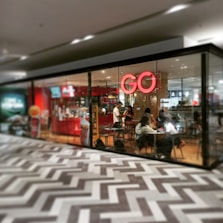 A glass-walled café with a prominent red neon sign that reads 'GO'. Inside, several people are seated at tables, engaged in conversations or working. The interior has a modern design with red and wooden elements, while the floor has a geometric zigzag pattern.