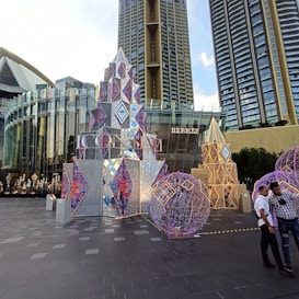 A modern urban environment with high-rise buildings in the background. The foreground features decorative structures including a geometric pyramid-like installation with vibrant patterns and colors, as well as spherical sculptures adorned with lights. The setting includes storefronts with visible brand signage.