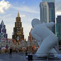 A large white inflatable sculpture is prominently featured in an outdoor setting surrounded by modern high-rise buildings. In the background, there are decorative structures with colorful geometric patterns. People are walking and interacting in the area, creating a lively atmosphere.