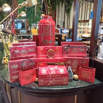 A festive display of red-themed tea products from TWG, featuring a large container labeled 'Red Christmas' surrounded by various red boxes and bags. The arrangement includes small decorative items like a miniature Christmas tree and gold ornaments, set on a dark green marble counter. Elegant brass lamps arch over the display, and the background includes greenery and a glimpse of a café setting.
