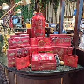 A festive display of red-themed tea products from TWG, featuring a large container labeled 'Red Christmas' surrounded by various red boxes and bags. The arrangement includes small decorative items like a miniature Christmas tree and gold ornaments, set on a dark green marble counter. Elegant brass lamps arch over the display, and the background includes greenery and a glimpse of a caf&eacute; setting.