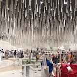 A modern shopping interior with a unique ceiling installation composed of numerous hanging cylindrical elements, creating a dynamic visual effect. The area is filled with various clothing racks, bags, and display cases filled with accessories. Shoppers can be seen browsing the items.