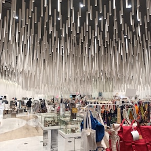 A modern shopping interior with a unique ceiling installation composed of numerous hanging cylindrical elements, creating a dynamic visual effect. The area is filled with various clothing racks, bags, and display cases filled with accessories. Shoppers can be seen browsing the items.