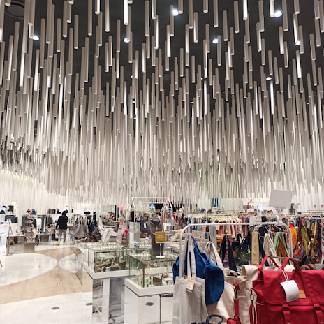 A modern shopping interior with a unique ceiling installation composed of numerous hanging cylindrical elements, creating a dynamic visual effect. The area is filled with various clothing racks, bags, and display cases filled with accessories. Shoppers can be seen browsing the items.
