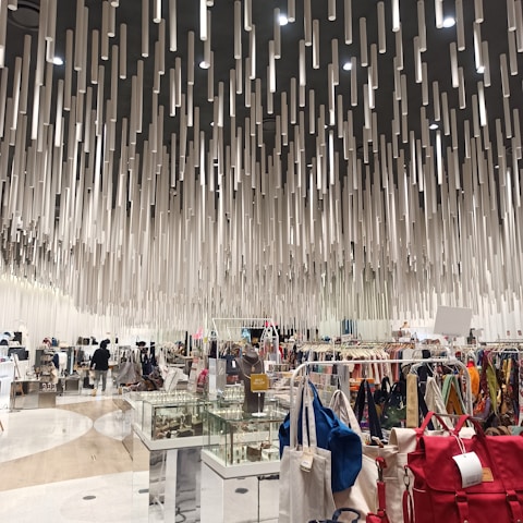 A modern shopping interior with a unique ceiling installation composed of numerous hanging cylindrical elements, creating a dynamic visual effect. The area is filled with various clothing racks, bags, and display cases filled with accessories. Shoppers can be seen browsing the items.