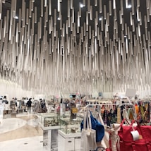 A modern shopping interior with a unique ceiling installation composed of numerous hanging cylindrical elements, creating a dynamic visual effect. The area is filled with various clothing racks, bags, and display cases filled with accessories. Shoppers can be seen browsing the items.