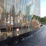 A modern architectural structure with a glass facade is adorned with intricate sculptures and decorative installations. These installations consist of multiple ornate designs in gold and silver hues, arranged elegantly along a reflective water feature. In the background, there are tall buildings and greenery, contributing to a serene urban setting.