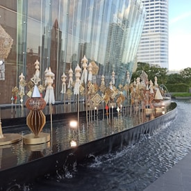 A modern architectural structure with a glass facade is adorned with intricate sculptures and decorative installations. These installations consist of multiple ornate designs in gold and silver hues, arranged elegantly along a reflective water feature. In the background, there are tall buildings and greenery, contributing to a serene urban setting.