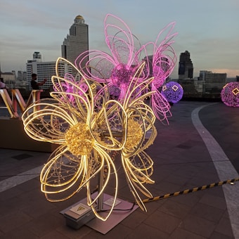 Brightly lit wire sculptures resembling flowers are arranged on a rooftop with a city skyline in the background. The sculptures are adorned with glowing yellow and pink lights, adding a vibrant contrast to the urban setting.