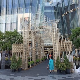 A decorative metal archway with intricate patterns leads to a shopping mall entrance. There is abundant greenery on either side, with several potted plants. The mall facade is modern, featuring glass panels and a sign that reads 'ICON SIAM'. Above, the names of luxury brands GUCCI and BOTTEGA VENETA are visible. A woman in a blue dress is walking toward the entrance.