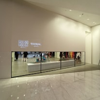 A minimalist store front with a large display window showcasing a modern and stylish interior. Two illuminated signs read 'PLEATS PLEASE' and 'BAO BAO ISSEY MIYAKE'. Several colorful geometric items are displayed on a long white counter. The floor is polished marble with a reflective surface.