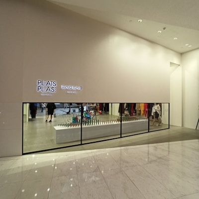 A minimalist store front with a large display window showcasing a modern and stylish interior. Two illuminated signs read 'PLEATS PLEASE' and 'BAO BAO ISSEY MIYAKE'. Several colorful geometric items are displayed on a long white counter. The floor is polished marble with a reflective surface.