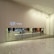 A minimalist store front with a large display window showcasing a modern and stylish interior. Two illuminated signs read 'PLEATS PLEASE' and 'BAO BAO ISSEY MIYAKE'. Several colorful geometric items are displayed on a long white counter. The floor is polished marble with a reflective surface.