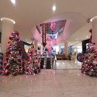 A festive shopping mall interior featuring elaborate Christmas decorations, including brightly colored Christmas trees adorned with oversized ornaments and candy-themed decorations. The area is illuminated with pink lights, and an escalator is visible in the center leading to an upper floor labeled as 'Gourmet Market'. Shoppers and seating areas are positioned in the background.