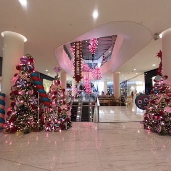 A festive shopping mall interior featuring elaborate Christmas decorations, including brightly colored Christmas trees adorned with oversized ornaments and candy-themed decorations. The area is illuminated with pink lights, and an escalator is visible in the center leading to an upper floor labeled as 'Gourmet Market'. Shoppers and seating areas are positioned in the background.