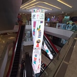 A shopping mall interior featuring an escalator and a tall, brightly lit pillar with colorful graphics and text. The pillar displays arrows, geometric patterns, and phrases like 'Enjoy' and 'Empower the Youth's Nextperience.' The surrounding area includes modern decor and ambient lighting.