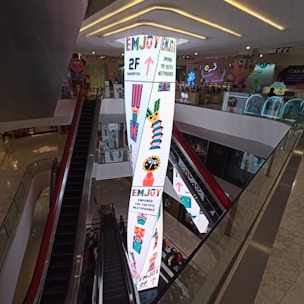 A shopping mall interior featuring an escalator and a tall, brightly lit pillar with colorful graphics and text. The pillar displays arrows, geometric patterns, and phrases like 'Enjoy' and 'Empower the Youth's Nextperience.' The surrounding area includes modern decor and ambient lighting.
