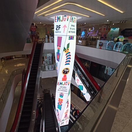 A shopping mall interior featuring an escalator and a tall, brightly lit pillar with colorful graphics and text. The pillar displays arrows, geometric patterns, and phrases like 'Enjoy' and 'Empower the Youth's Nextperience.' The surrounding area includes modern decor and ambient lighting.