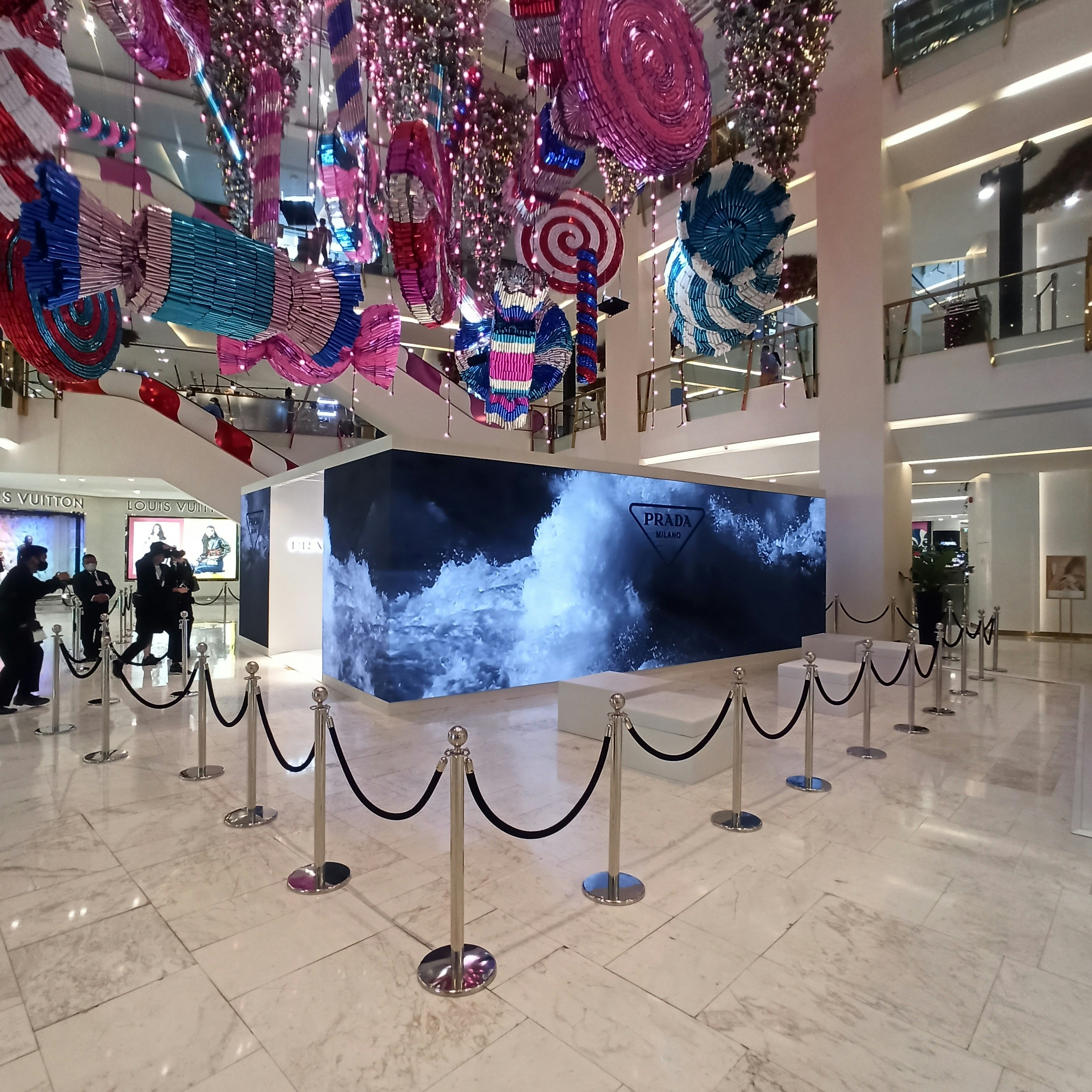 A retail store interior features a large Prada display surrounded by stanchions, with decorative, oversized candy-like ornaments hanging from the ceiling in vibrant colors. The floor is polished white marble, and the store has high ceilings with multiple floors visible. People are seen in the background near other luxury brand displays.