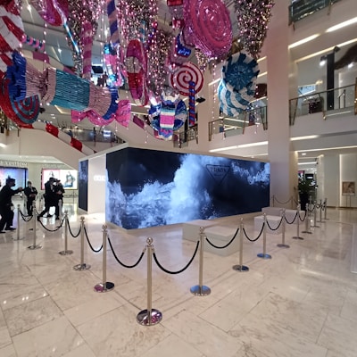 A retail store interior features a large Prada display surrounded by stanchions, with decorative, oversized candy-like ornaments hanging from the ceiling in vibrant colors. The floor is polished white marble, and the store has high ceilings with multiple floors visible. People are seen in the background near other luxury brand displays.