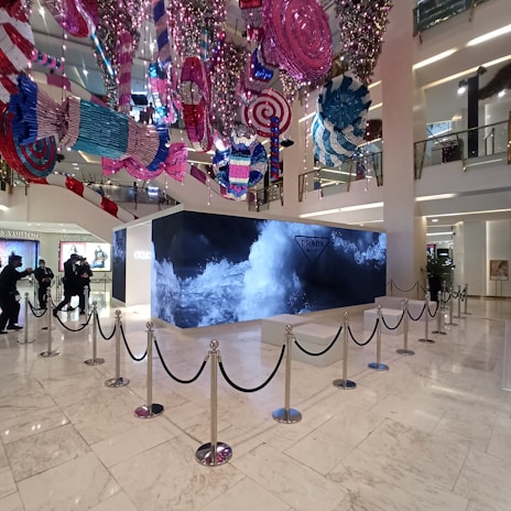 A retail store interior features a large Prada display surrounded by stanchions, with decorative, oversized candy-like ornaments hanging from the ceiling in vibrant colors. The floor is polished white marble, and the store has high ceilings with multiple floors visible. People are seen in the background near other luxury brand displays.