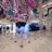 A retail store interior features a large Prada display surrounded by stanchions, with decorative, oversized candy-like ornaments hanging from the ceiling in vibrant colors. The floor is polished white marble, and the store has high ceilings with multiple floors visible. People are seen in the background near other luxury brand displays.